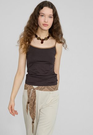 Junge Frau mit langen lockigen Haaren, die ein schwarzes Tanktop, beige Hose, einen gemusterten Schärpengürtel und eine breite braune Halskette trägt, vor einem schlichten Hintergrund stehend.