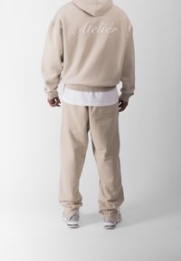 Sudadera beige con "Atelier" bordado en la parte posterior, combinada con pantalones de chándal a juego. Textura suave, corte relajado, puños y cintura acanalados.