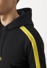 Hoodie noir avec un accent rayé jaune le long de l'épaule et un branding texturé. Comprend une capuche ajustable avec cordons.