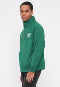 Temno zelena pulover s četrt zadrgo in stojnim ovratnikom, okrašen z belim logotipom "Calvin Klein", z flisasto teksturo ter rebrastimi manšetami in robom.