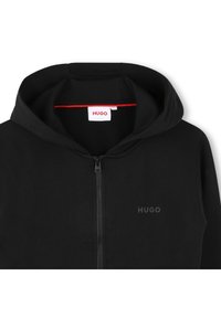 Hoodie zippé noir en tissu texturé, avec capuche, fermeture éclair frontale et un petit logo noir au-dessus de la fermeture. Accent rouge à l'intérieur de la capuche.