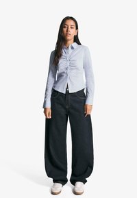 Striped button-up shirt in light blue with gathers at the front, paired with oversized dark denim trousers and white sneakers.
