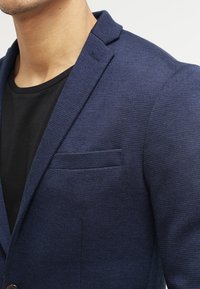 Marinblå texturerad blazer med slagkrage, enkel bröstdesign och bröstficka, bärs över en svart skjorta.