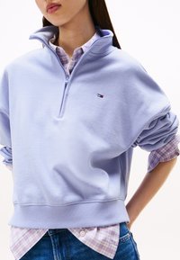 Sudadera de color lila claro con zip de cuarto y ribete en el dobladillo y en los puños, llevada sobre una camisa a cuadros. Presenta un pequeño logo en el pecho. Ajuste casual.