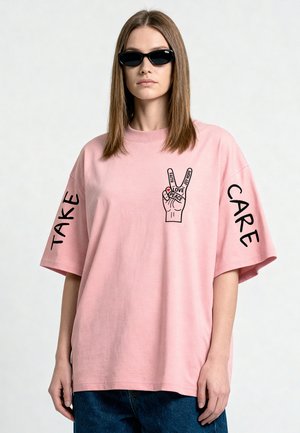 Vrouw met zwarte zonnebril en een roze oversized T-shirt met vredesteken en de woorden "TAKE CARE" op de mouwen.