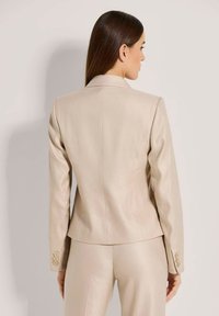 Beige op maat gemaakte blazer met notch-kleppen, getailleerde silhouet en drieknoopmanchetten. Soepele textuur met subtiele glans.