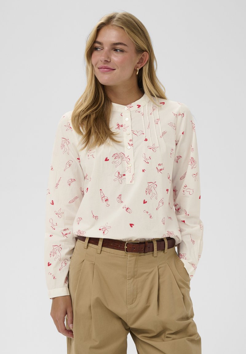 Krémová blúzka s dlhým rukávom s červenými fantazijnými kresbami, s gombíkmi pri výstrihu v štýle henley a vo voľnom strihu. Kombinovaná s béžovými nohavicami.