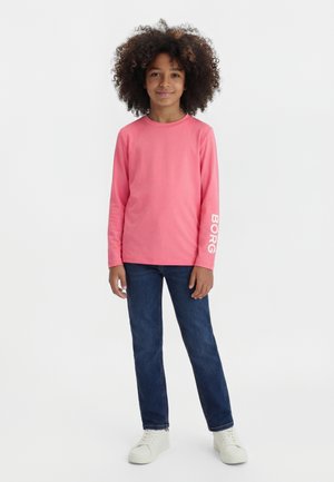 Enfant aux cheveux bouclés portant un t-shirt rose à manches longues avec « BORG » sur la manche, un jean bleu foncé et des baskets blanches, debout face à l'avant.