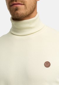 Pull en maille côtelée de couleur crème avec un patch logo circulaire marron sur le côté gauche de la poitrine. Texture douce et coupe ajustée.