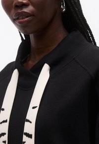 Close-up af en person iført sort hættetrøje med tykke hvide snore med sorte broderede detaljer, og et lille hult øreørering synlig.