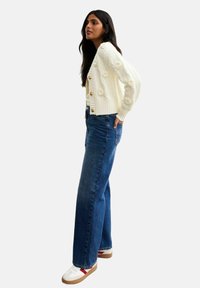 Cardigan in maglia crema con design floreali a texture e bottoni in legno, abbinato a jeans blu a vita alta e sneakers bianche con dettagli rossi.