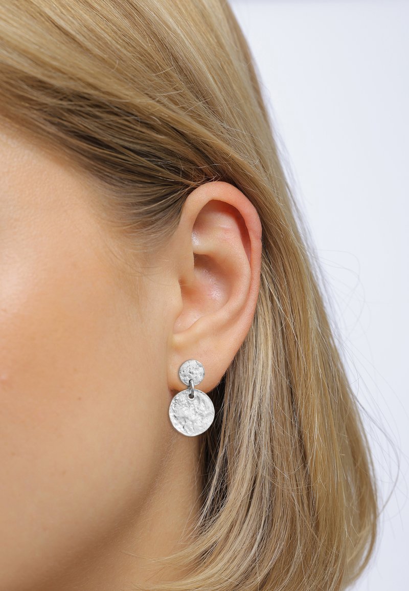Silberne Ohrringe mit einem strukturierten runden Anhänger und einem kleineren runden Stecker, verbunden durch einen Metallring, am Ohrläppchen getragen.