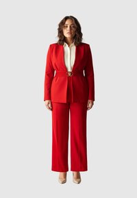 Roter maßgeschneiderter Anzug mit tailliertem Blazer, weitem Hosenbein, goldenem Gürtelverschluss und weißem Hemd darunter. Glatter Stoff, strukturierte Gestaltung.