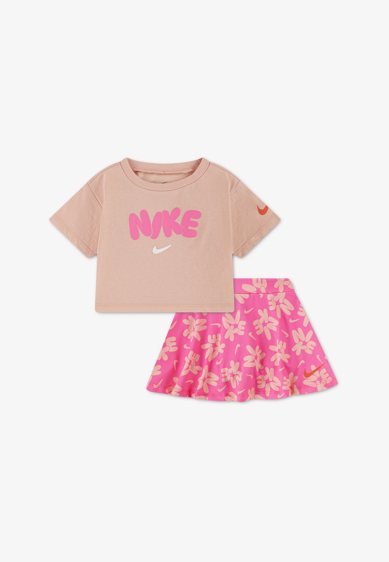 T-shirt beige court avec texte "NIKE" rose et virgule blanche, assorti à une jupe rose avec motifs floraux beiges et virgule.