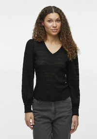 Svart stickad tröja med V-hals, långa puffärmar och texturerad detaljering genom hela tyget, kombinerad med grå jeans.