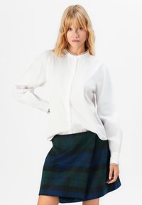 Kvinne med blondt hår iført en hvit knappebluse med blonder, poserer med hånden på hoften, iført et rutete skjørt i grønn og blå.