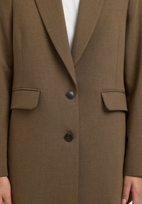 Hnědý sako na míru s klopami s výřezem, dvěma černými knoflíky, kapsami s klopami, nošený přes bílou košili.