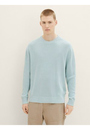 Hellblauer gestrickter Pullover mit Rundhalsausschnitt, gerippten Bündchen und Saum, der ein strukturiertes Muster aufweist. Wird mit beigen Hosen getragen.