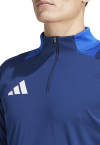 Haut demi-zip de sport marine avec des accents bleus, fabriqué en tissu lisse et extensible. Présente un logo blanc sur la poitrine et un col zippé.