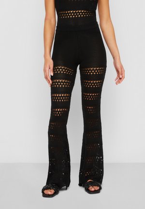 Woman wearing black crochet knit flare pants with patterned openwork and black braided slide sandals.