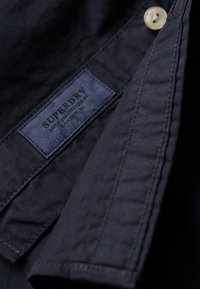 Marinekleurige overhemdkraag met een label "SUPERDRY." De stof is glad, met een knoopdetail bij de hals. Minimale stiknaden zichtbaar.