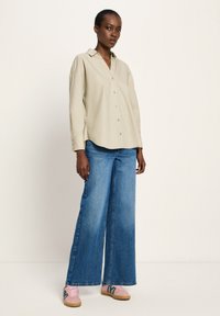 Woman wearing a beige button-up long-sleeve shirt, wide-leg blue jeans, and pink sneakers standing against a plain light background.
