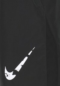 Černé sportovní šortky s velkým, opotřebovaným bílým logem Nike swoosh na levé straně, se viditelnými detaily šití.