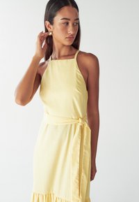 Geel mouwloos jurkje van gestructureerde stof, met een strik taille en een ruchezoom, dunne bandjes en een eenvoudig, getailleerd ontwerp.