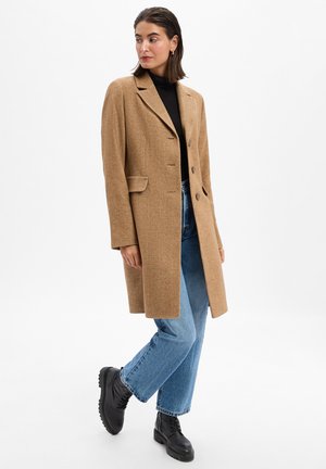 Brown herringbone coat with a structured design, two buttons, notched lapels, and front pockets, paired with wide-leg blue jeans and black boots.