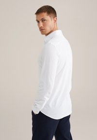 Fehér, hosszú ujjú galléros póló, gombos elülső résszel és testhez álló szabással. Sötétkék nadrággal viselve, sima textúrával.