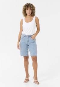 Pantaloni shorts in denim azzurro chiaro con orli arrotolati, cinque tasche e una vestibilità rilassata, abbinati a una canotta bianca attillata e sandali con tacco trasparente.