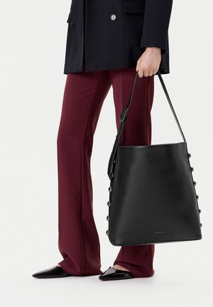 Persona con pantalones burdeos y chaqueta negra sostiene un gran bolso de cuero negro con tachuelas en los lados y una correa para el hombro.