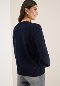Navyblauwe gebreide trui met lange mouwen, ronde halslijn en ribgebreide zoom. Zachte textuur met subtiele glans. Model staat met de rug naar de camera en toont de details aan de achterkant.