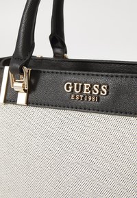 Bolso de mano de imitación de cuero negro con herrajes dorados, que presenta un cuerpo de tela blanca con un patrón texturizado. Logo "GUESS EST 1981" mostrado de manera prominente.