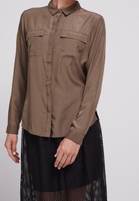 Camicia marrone a maniche lunghe in materiale lucido, con colletto e due tasche sul petto. Abbinata a una gonna in rete nera, che mette in mostra texture sovrapposte.