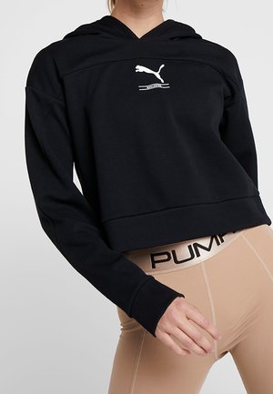 Osoba nosząca czarną krótką bluzę z kapturem Puma i beżowe legginsy z widocznym paskiem z logo Puma na jednolitym tle.