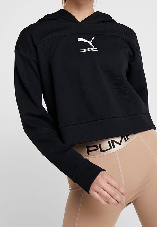 Personne portant un sweat à capuche court noir Puma et des leggings beige avec une ceinture visible au logo Puma sur un fond uni.