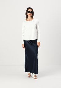 White long-sleeve top, soft texture, paired with a flowing navy satin maxi skirt. Model wears large black sunglasses and white strappy heels.