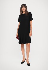 Black short-sleeve dress with a flared hem, made of soft material. Paired with black pointed-toe heels and a red clutch.