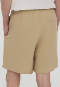 Pantaloni corti beige con cintura elastica, tasca posteriore e texture liscia. L'orlo è dritto e termina sopra il ginocchio.