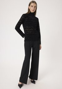 Schwarzes Spitzenoberteil mit hohem Kragen und langen Ärmeln, kombiniert mit hochgeschnittenen schwarzen Hosen und spitzen schwarzen Absätzen. Glatte Textur und kompliziertes Muster.