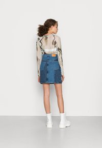 Saia de ganga azul com cintura ajustada, dois bolsos traseiros e detalhe em bainha desgastada. Combinada com uma blusa transparente de mangas longas e padrão, e botas brancas.