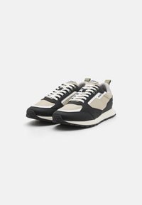 Des baskets mélangeant des matériaux noirs, beiges et vert olive, avec une surface texturée, des lacets blancs et un design simple.
