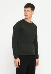 Camicia a maniche lunghe di un verde scuro, con texture, scollo rotondo, forma aderente e orlo a coste. Indossata con pantaloni neri.