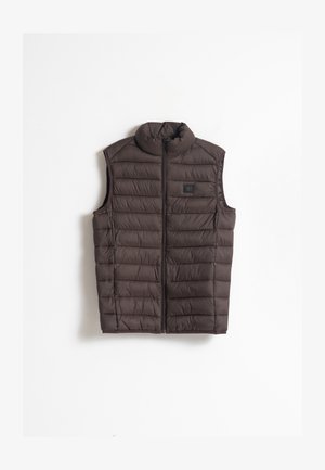 Chaleco sin mangas acolchado marrón con un cuello alto, cierre frontal y un pequeño parche de logo en la parte izquierda del pecho. Textura de tela ligera.