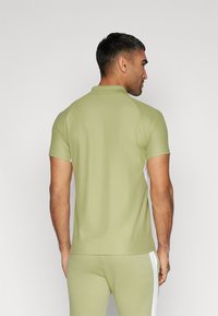Grøn polo-shirt med en diskret struktur, korte ærmer og kravehals. Har hvide sidekanter for kontrast. Vist fra bageste vinkel.