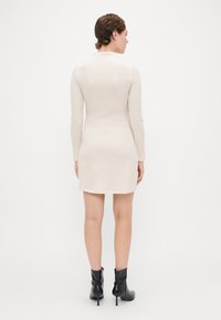Robe en tricot côtelé beige avec des manches longues et une coupe ajustée, se terminant au milieu de la cuisse. Portée avec des bottines noires, texture lisse, design minimal.