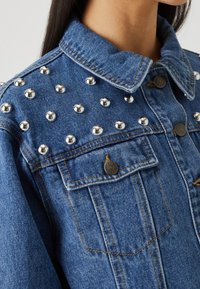 Farkkutakki hopeisilla niiteillä olkapäissä, jossa on klassinen kaulus, napitettu rintatasku ja kontrastivärinenommeltu.