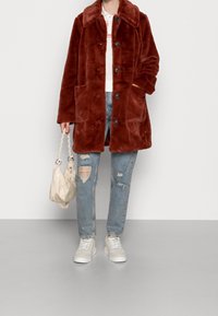 Person iført en rustfarvet faux fur jakke, slidte blå jeans, hvide sneakers og holder en cremet håndtaske med flet-stropper.
