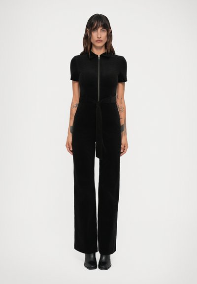 Vrouw die naar voren staat in een zwarte short-sleeve jumpsuit met rits aan de voorkant, riem en zwarte enkellaarsjes op een plain witte achtergrond.
